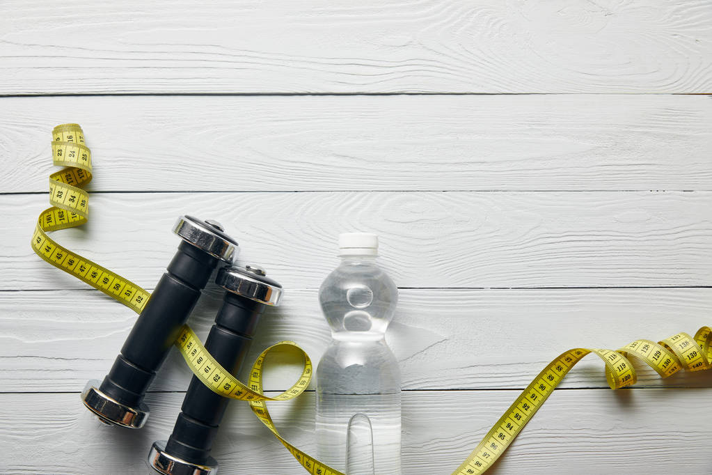 vista superior de la botella con agua, cinta métrica amarilla y mancuernas sobre fondo blanco de madera con espacio para copiar
 - Foto, Imagen