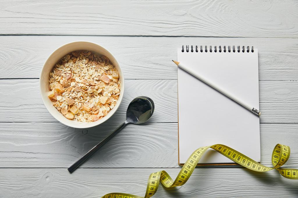 vista superior de fita métrica, notebook, lápis, colher e cereal café da manhã em tigela sobre fundo branco de madeira
  - Foto, Imagem