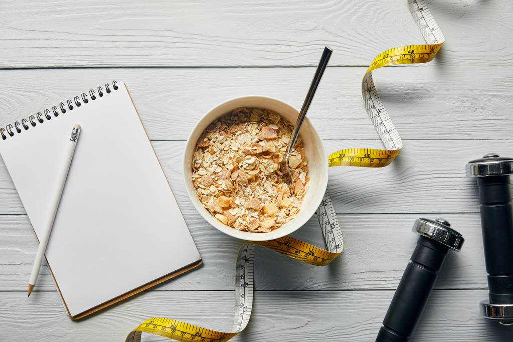 top view of measuring tape, spoon and breakfast cereal in bowl near notebook, dumbbells and pencil on wooden white background  - Photo, Image