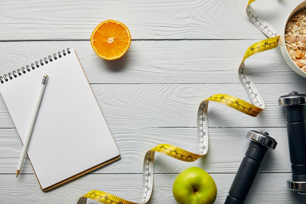 верхний вид измерительной ленты, хлопья на завтрак в миске рядом с яблоком, апельсин, ноутбук, гантели и карандаш на деревянном белом фоне с копировальным пространством
 - Фото, изображение