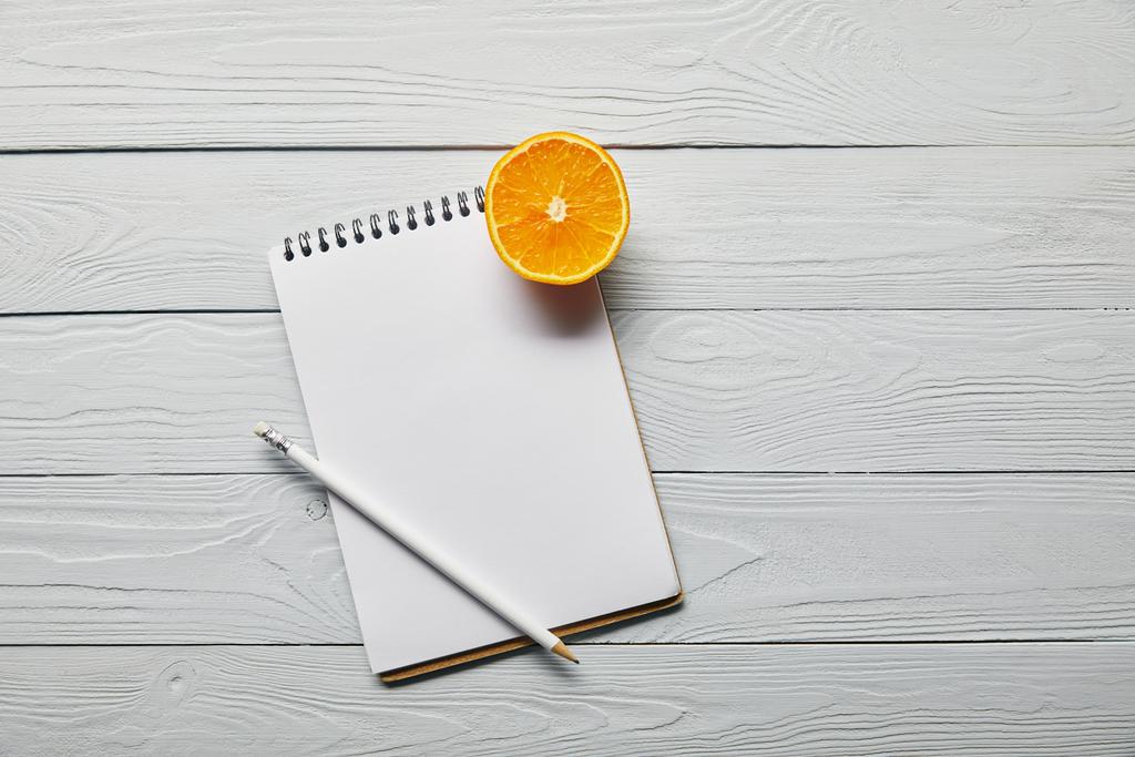pohled na oranžovou půlku, prázdný Poznámkový blok a tužku na dřevěné bílé pozadí s prostorem pro kopírování - Fotografie, Obrázek