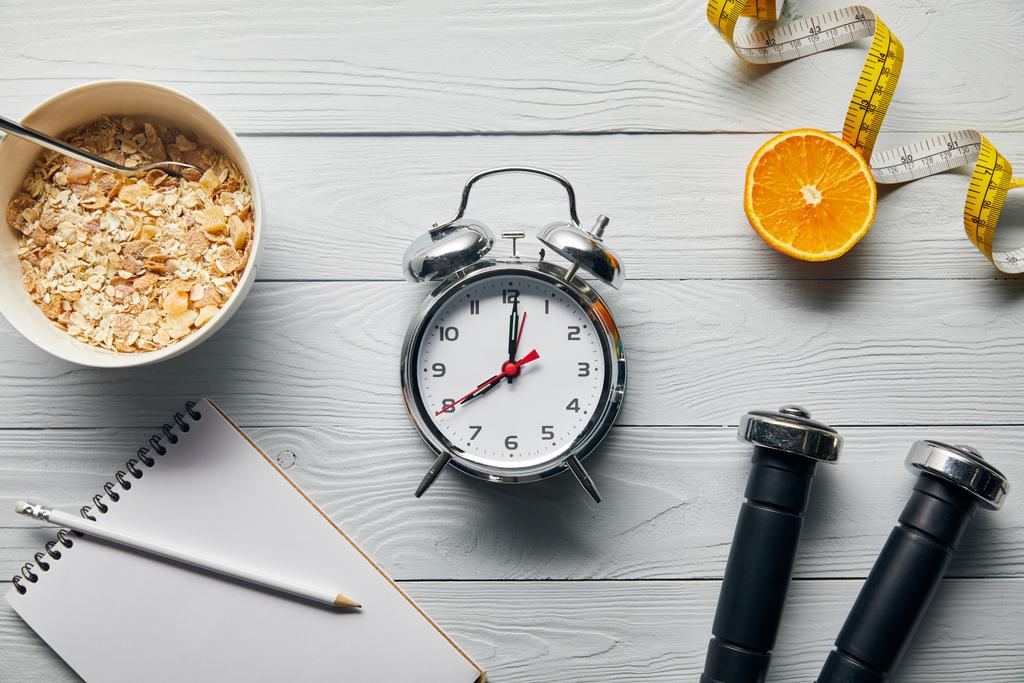 top view of silver alarm clock, notebook with pencil, breakfast cereal, measuring tape, dumbbells and orange half on wooden white background  - Photo, Image