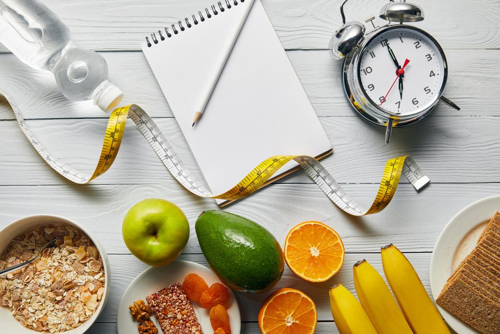 Top uitzicht van dieet voedsel in de buurt van wekker, meetlint, notebook en water op houten witte achtergrond  - Foto, afbeelding