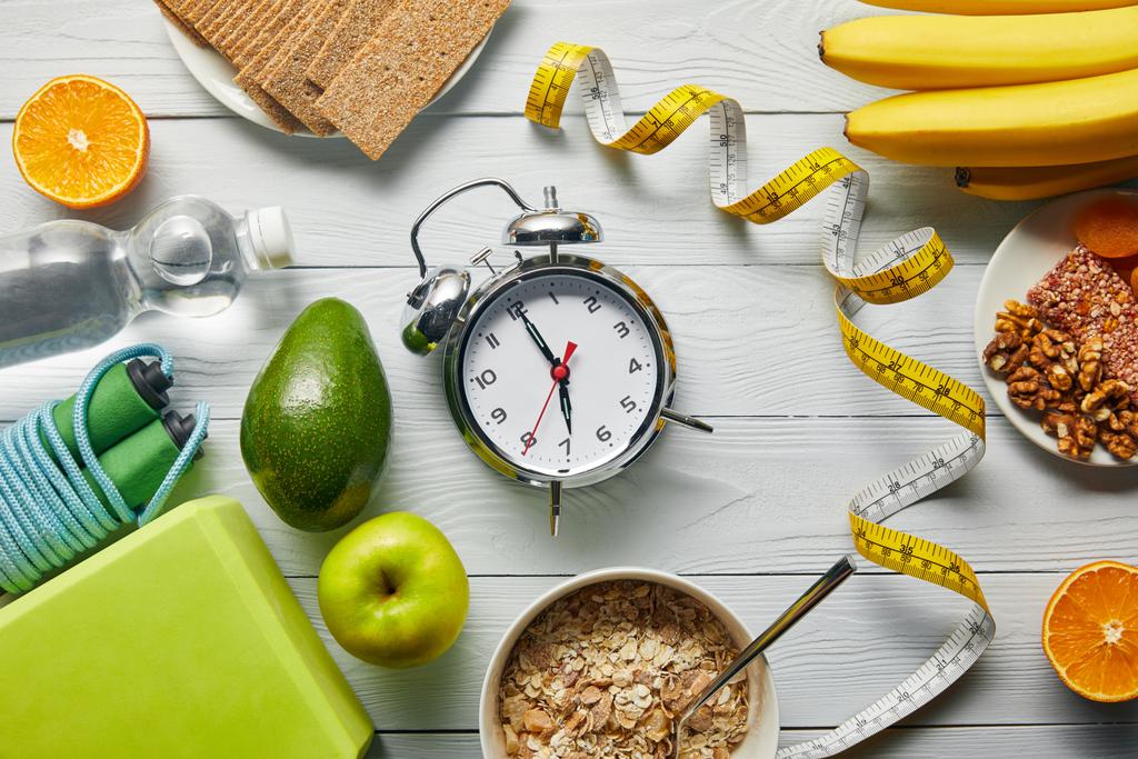 верхний вид диетической пищи возле измерительной ленты, будильника и спортивного оборудования на деревянном белом фоне
 - Фото, изображение