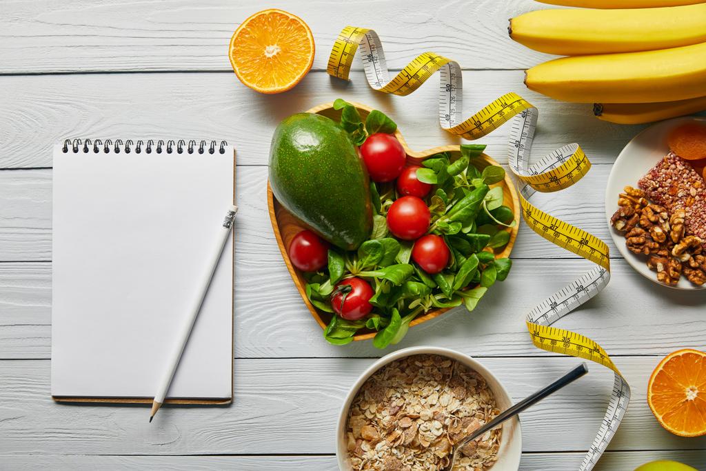 Maßband, leeres Notizbuch und Müsli, Obst und Gemüse auf weißem Holzgrund  - Foto, Bild