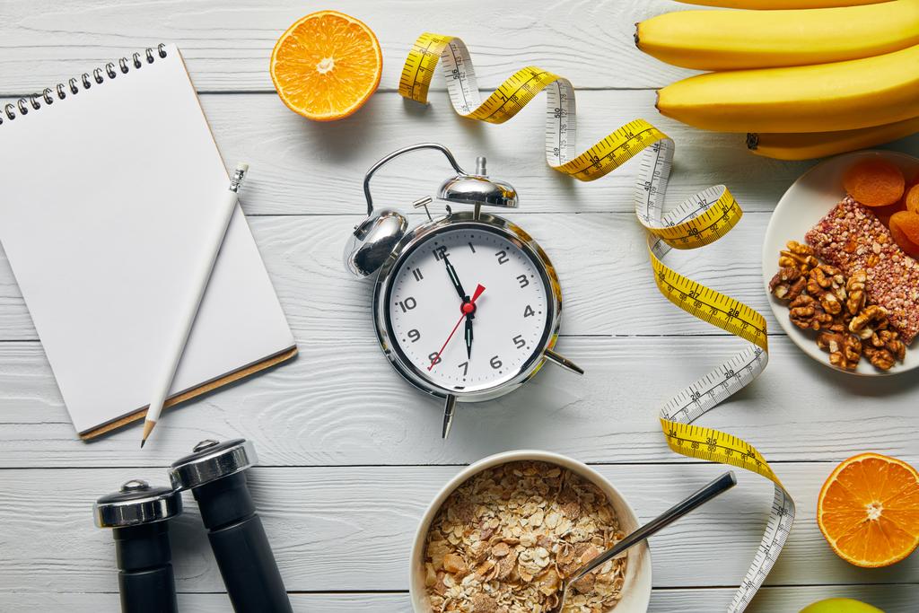 vista superior de cinta métrica, comida dietética y pesas cerca del cuaderno en blanco y reloj despertador sobre fondo blanco de madera
  - Foto, Imagen