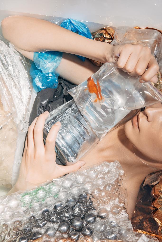 обрезанный вид мокрой женщины, держащей золотую рыбку в пластиковом пакете среди мусора в ванной
 - Фото, изображение