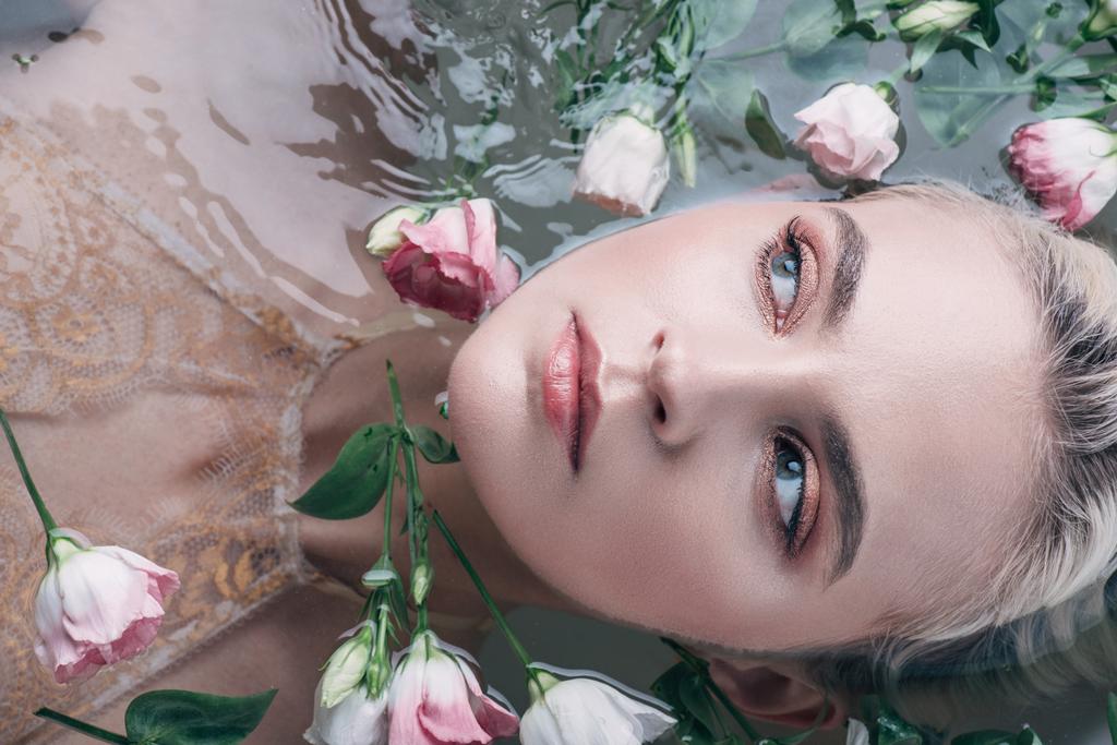 Draufsicht der schönen Frau, die im klaren Wasser zwischen Blumen liegt und wegschaut - Foto, Bild