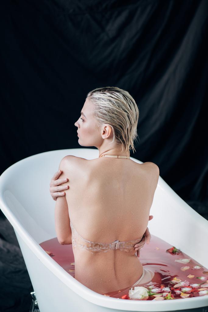 Rückansicht der blonden sexy nassen Frau, die in einem Bad mit rosa Wasser und Rosenblättern sitzt - Foto, Bild