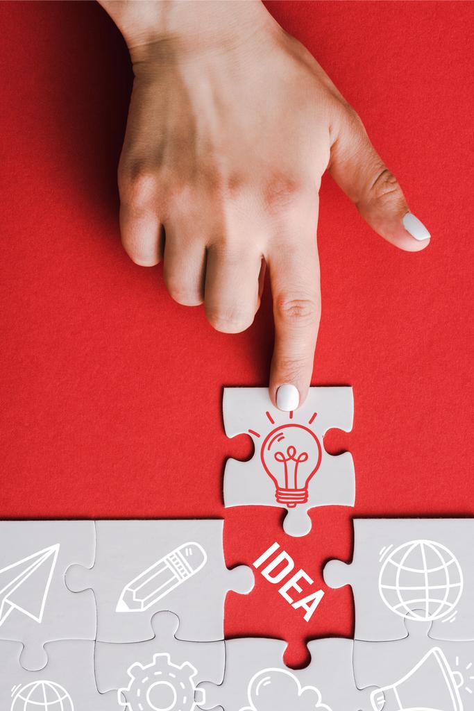 top view of woman pointing with finger at jigsaw with light bulb near connected white puzzle pieces and idea lettering on red - Photo, Image
