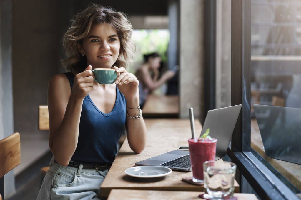 Очаровательная молодая успешная женщина внештатный менеджер Smm сидя журнальный столик выглядят мечтательно через окно прохожих держать чашку кофе улыбаясь с удовольствием взять перерыв рабочий ноутбук, делать исследования
 - Фото, изображение