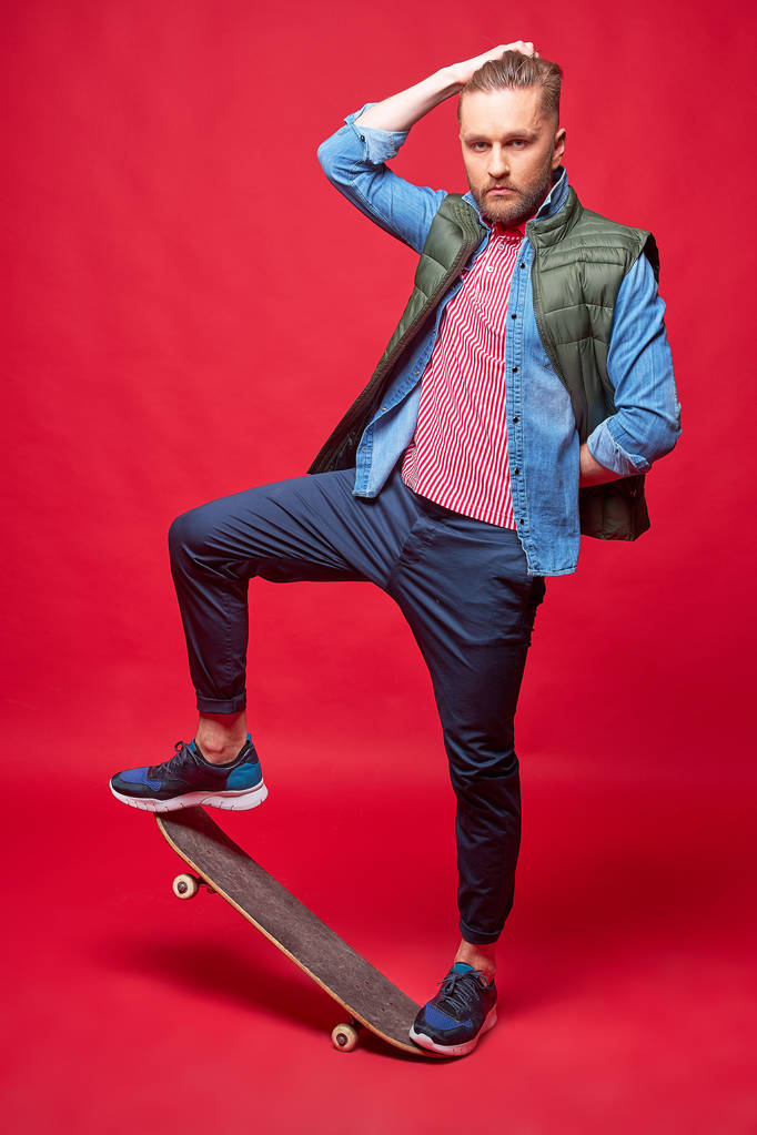 スケートボードに立つカジュアルなスタイルのファッショナブルな男。スタジオショット、レッドバックグラウンド. - 写真・画像