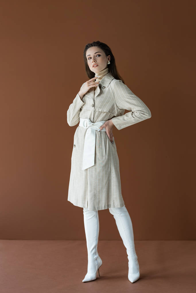 stylisches Modell im beigen Trenchcoat stehend mit Hand an der Hüfte isoliert auf braun - Foto, Bild