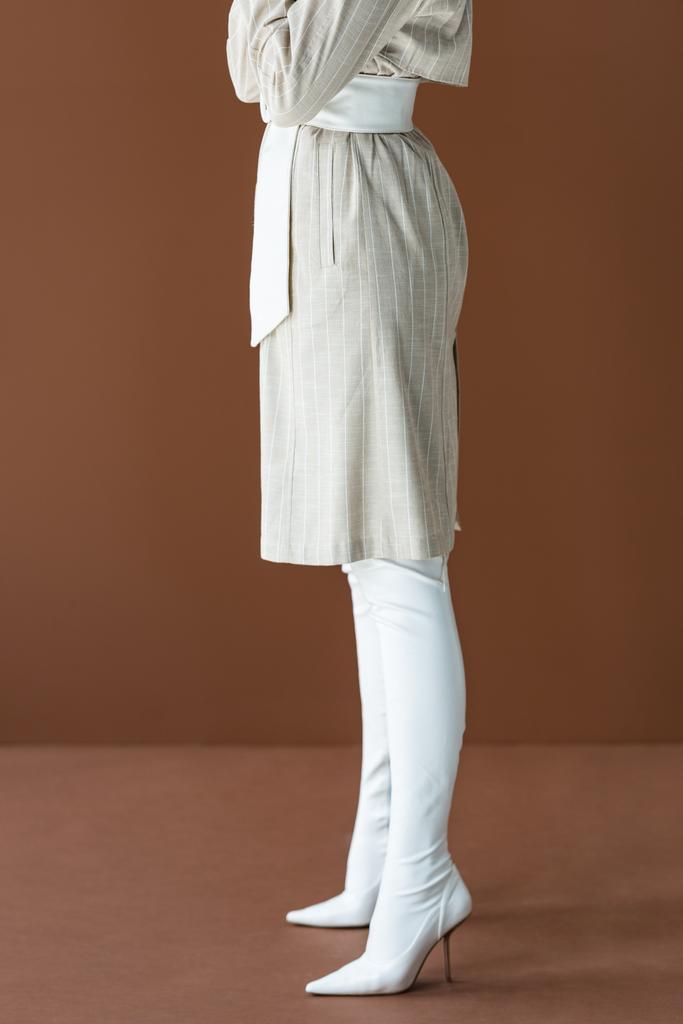 обрезанный вид на стильную женщину в плаще и белых сапогах, стоящих изолированы на коричневый
 - Фото, изображение