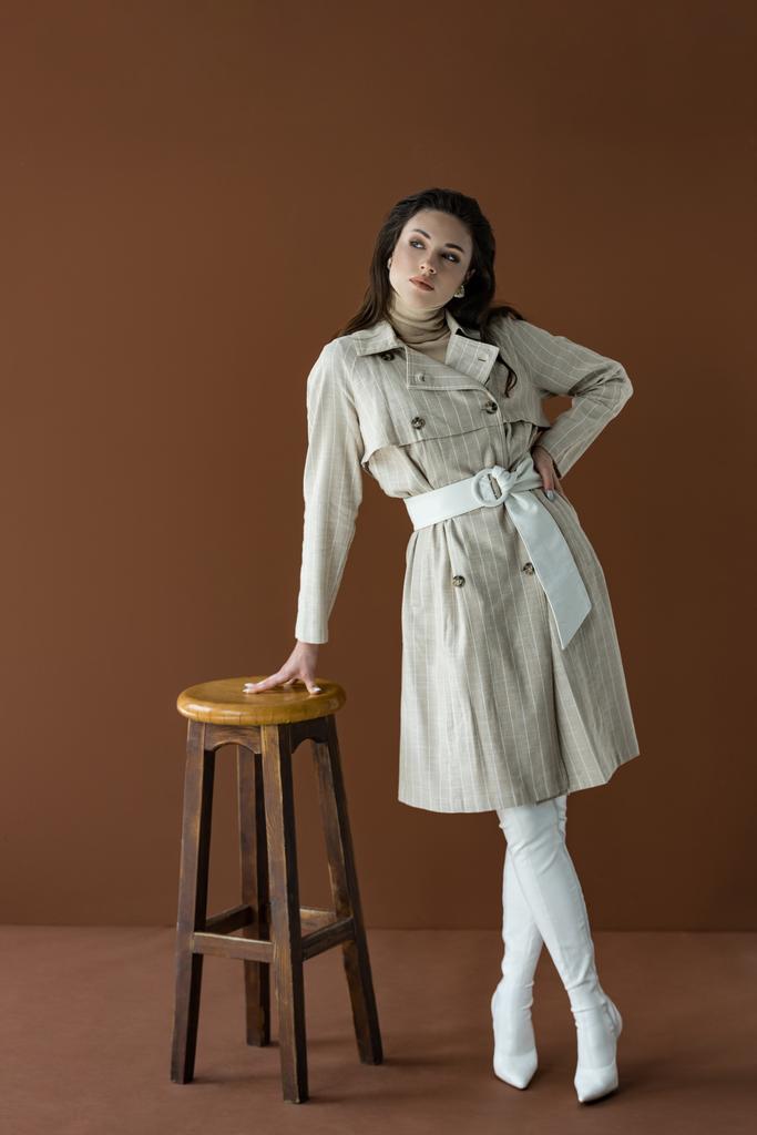 μοντέρνα γυναίκα σε κομψό καμπαρντίνα κοιτάζοντας μακριά, στέκεται κοντά καρέκλα απομονωμένη σε καφέ - Φωτογραφία, εικόνα