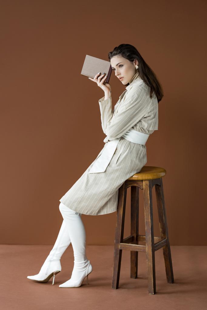 Seitenansicht der schönen stilvollen Frau im Trenchcoat mit Buch, sitzend auf Stuhl isoliert auf braun - Foto, Bild