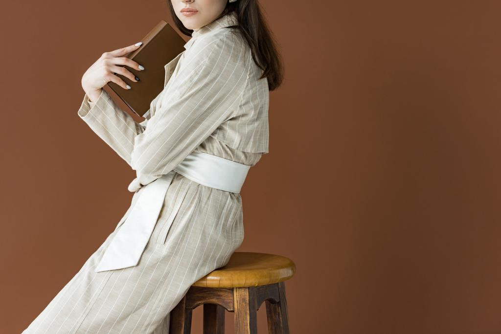 Ausgeschnittene Ansicht eines jungen Models, das Buch in der Hand hält, isoliert auf braun sitzend - Foto, Bild