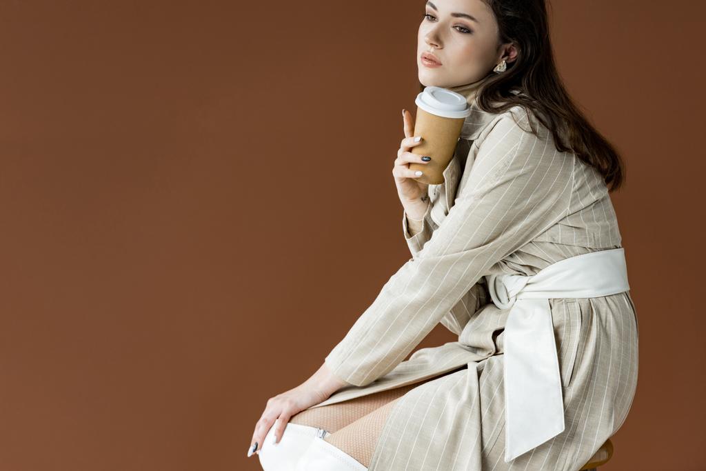 hermoso modelo mirando hacia otro lado, sentado aislado en marrón, sosteniendo la taza de papel en la mano
 - Foto, Imagen