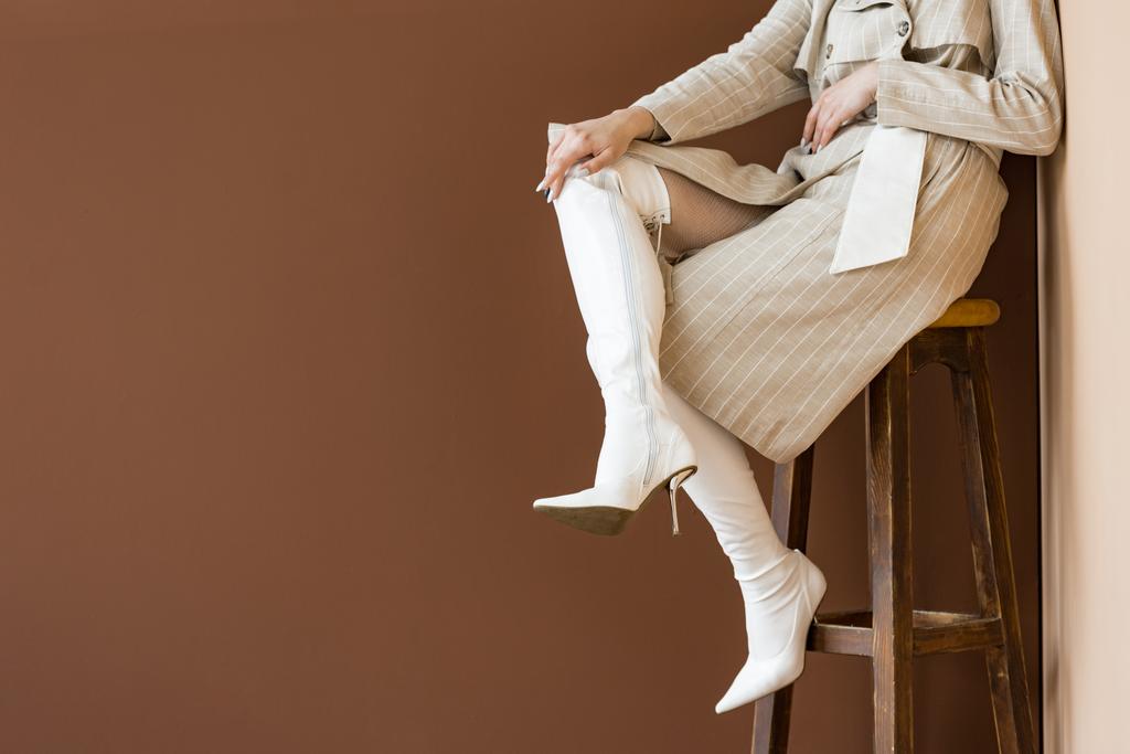 обрізаний вид на дорослу модель в бежевому траншеї пальто і білі черевики, що сидять на дерев'яному стільці
 - Фото, зображення