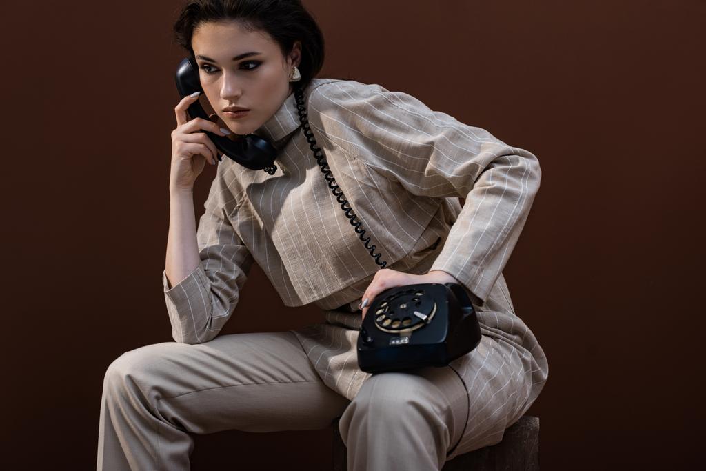modny model z słuchawką w ręku siedzi na brązowym tle, patrząc z dala - Zdjęcie, obraz