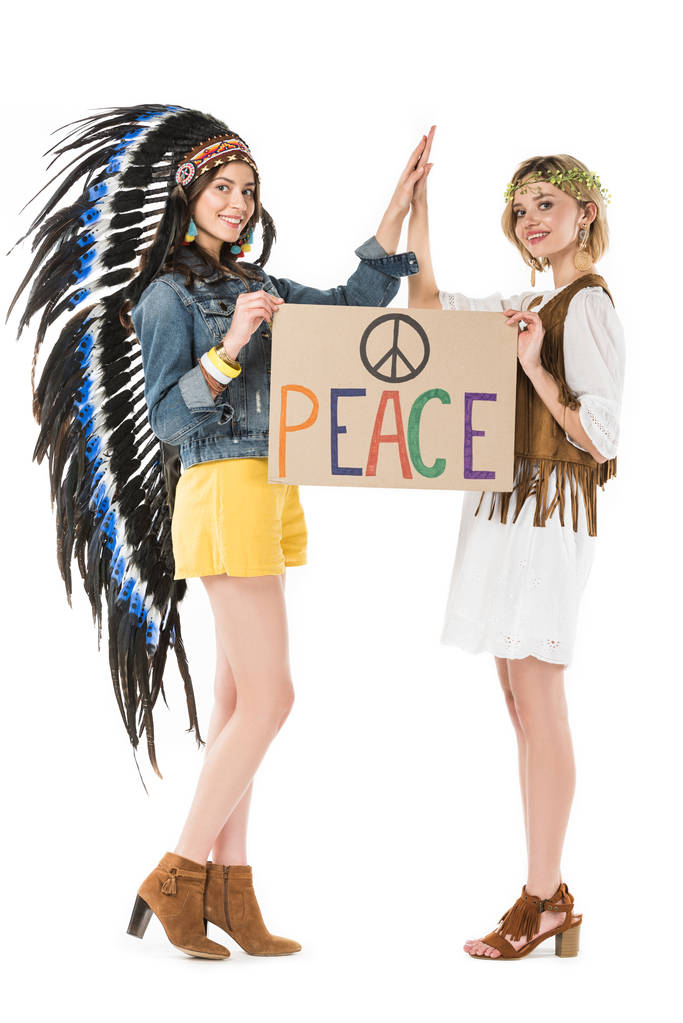 volledige lengte weergave van twee biseksuele hippie meisjes in Indiase hoofdtooi en krans houden plakkaat met inscriptie en aanraken van handen geïsoleerd op wit - Foto, afbeelding