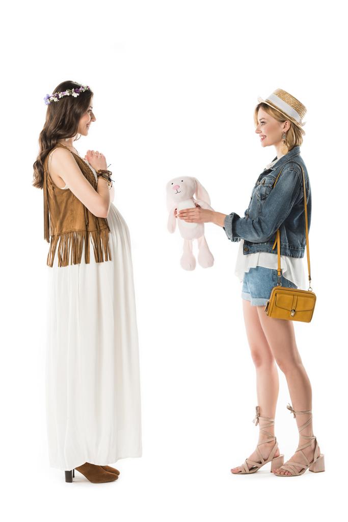 Zijaanzicht van hippie meisje geven speelgoed konijn aan zwangere vriendin geïsoleerd op wit - Foto, afbeelding