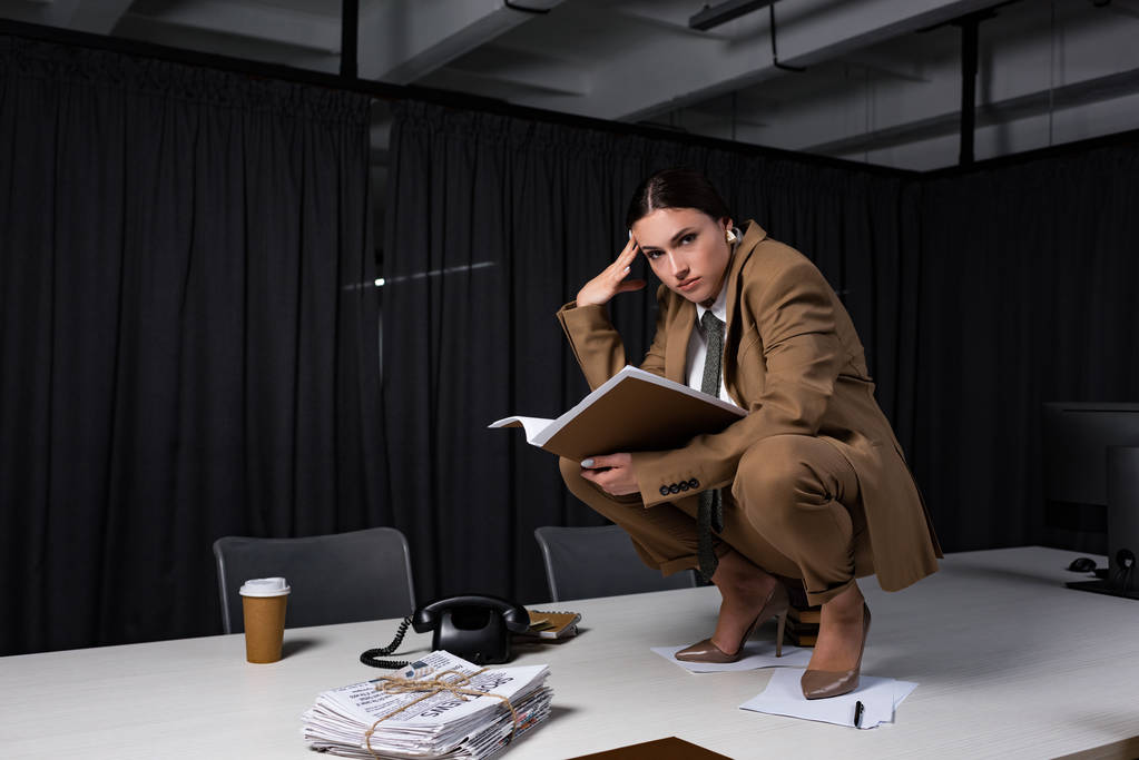 Stilvolle Geschäftsfrau im Anzug sitzt am Tisch, hält Dokumente in der Hand und blickt in die Kamera - Foto, Bild