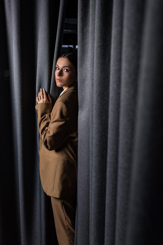 vista lateral da mulher bem sucedida em terno em pé sobre fundo de cortina cinza escuro, olhando para a câmera
 - Foto, Imagem