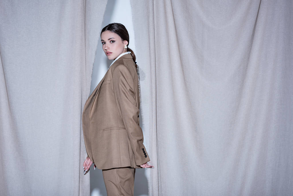 side view of successful woman in suit standing on light grey curtain background, looking at camera - Photo, Image