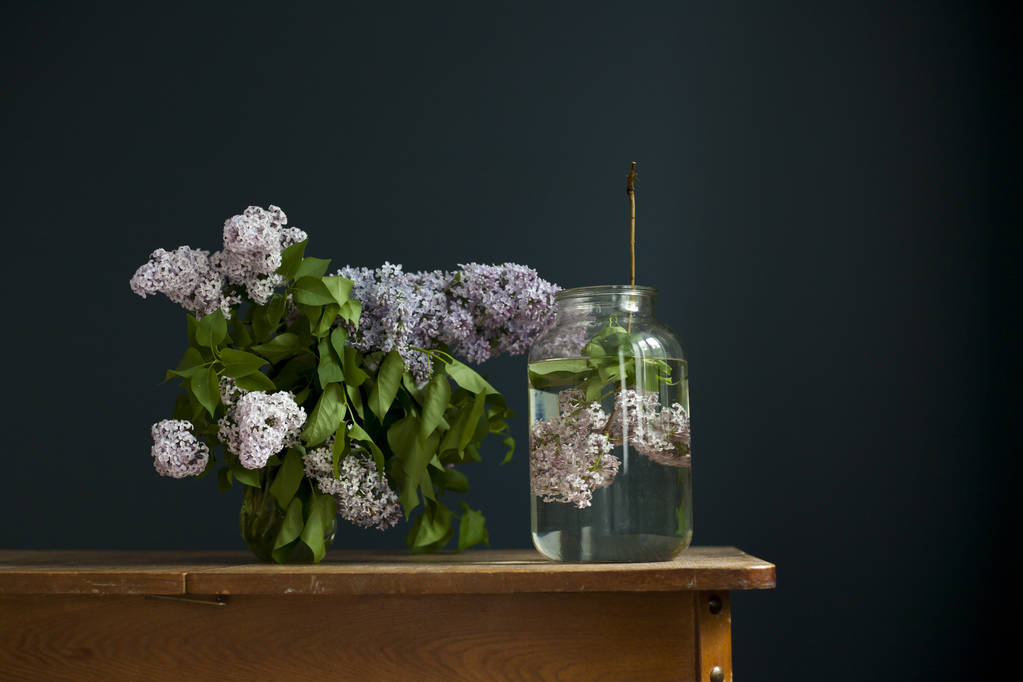 kytice fialových větviček v průhledné zelené nádobě na dřevěném stole naproti černé stěně - Fotografie, Obrázek