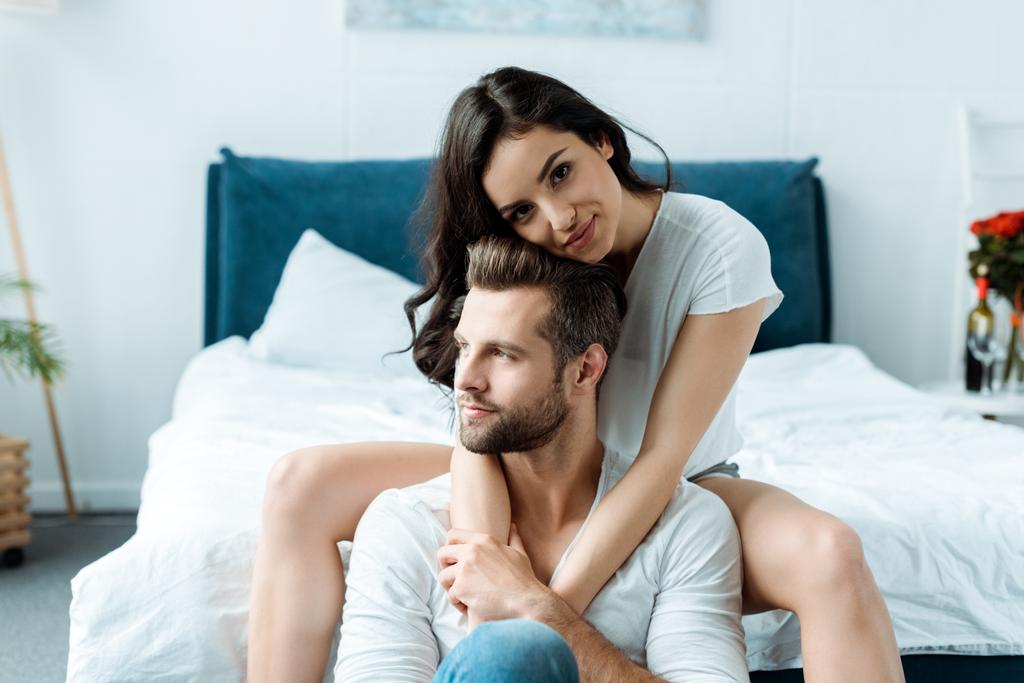 beautiful smiling woman hugging boyfriend and looking at camera in bedroom - Photo, Image