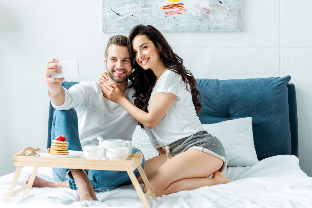 szczęśliwa para przytulanie i robienia selfie na smartfonie w pobliżu drewnianej tacy ze śniadaniem w łóżku - Zdjęcie, obraz