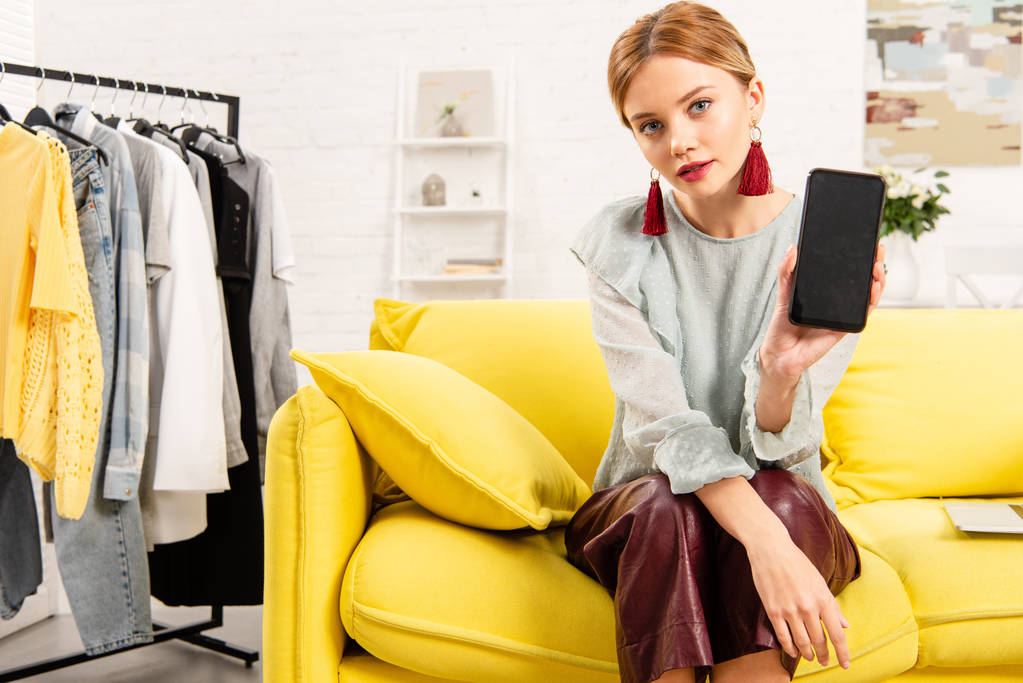 viehättävä tyttö trendikkäissä korvakoruissa istuu keltaisella sohvalla ja käyttää älypuhelinta kotona
 - Valokuva, kuva