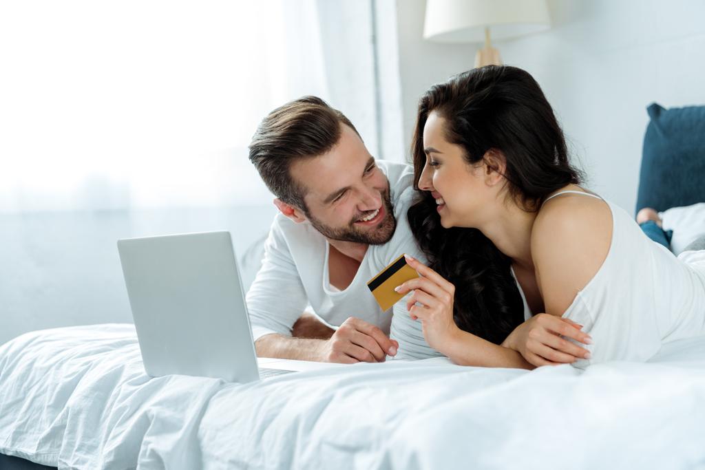 szczęśliwa para leżącego w łóżku w pobliżu laptopa, podczas gdy kobieta trzyma kartę kredytową, ilustrujący redakcyjnej - Zdjęcie, obraz