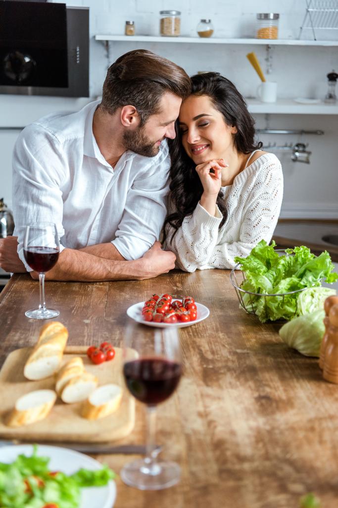 coppia tenera e sognante vicino al tavolo di legno con vino rosso, pomodorini, pane e insalata
 - Foto, immagini