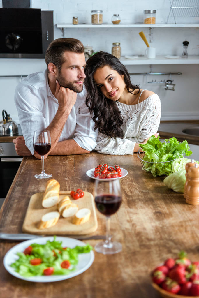couple souriant de rêve près de la table en bois avec vin rouge, tomates cerises, pain et salade
 - Photo, image