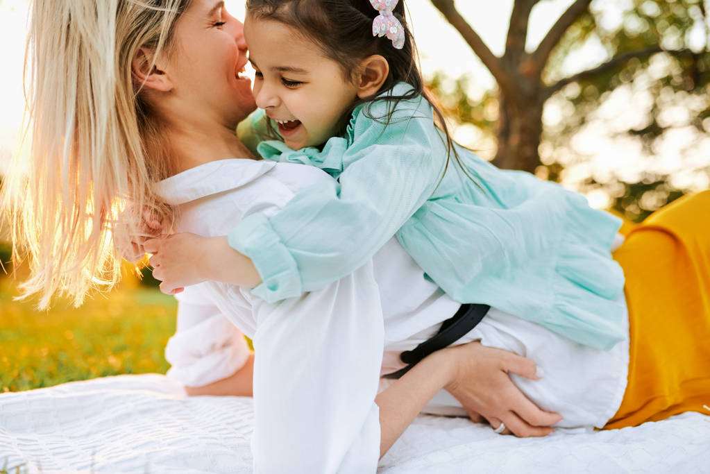 Εικόνα της ευτυχΙάς κόρης που χαμογελάει και αγκαλιάζει τη μητέρα της, περνώντας χρόνο μαζί στο πάρκο. Νεαρή γυναίκα που παίζει και διασκεδάζει με το παιδί της στο πράσινο γρασίδι. Η μαμά και το κοριτσάκι μοιράζονται την αγάπη. - Φωτογραφία, εικόνα