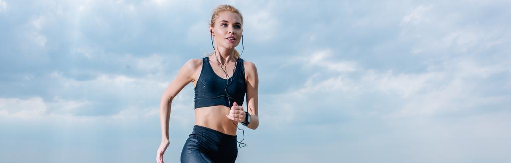 panoramic shot of sportive blonde woman running and listening music in earphones  - Photo, Image