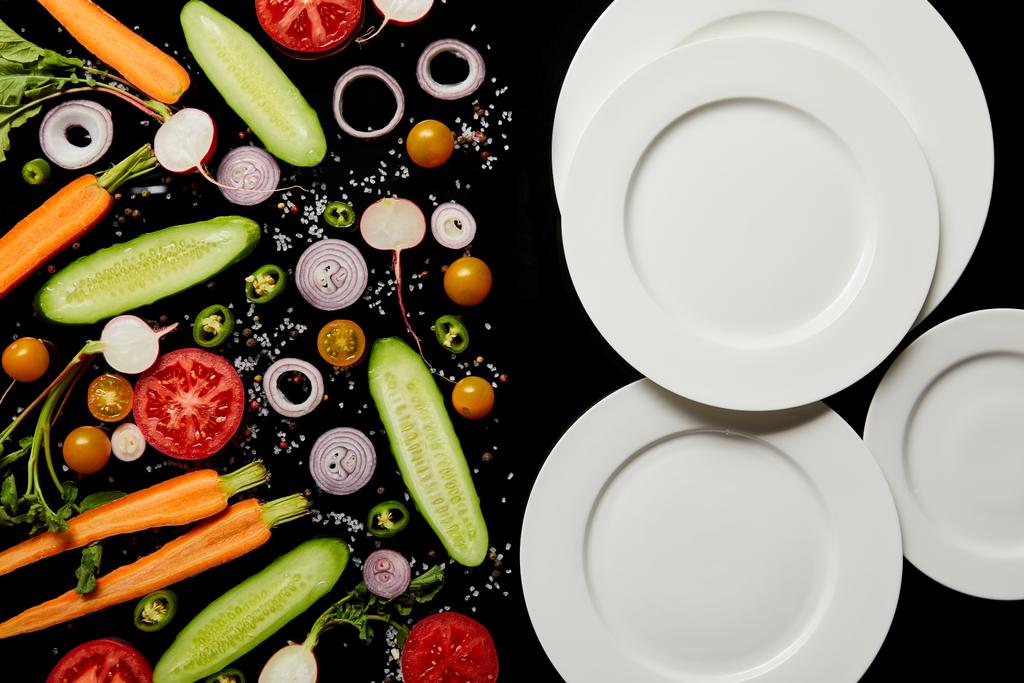 vista superior de platos redondos vacíos cerca de verduras en rodajas aisladas en negro
 - Foto, Imagen