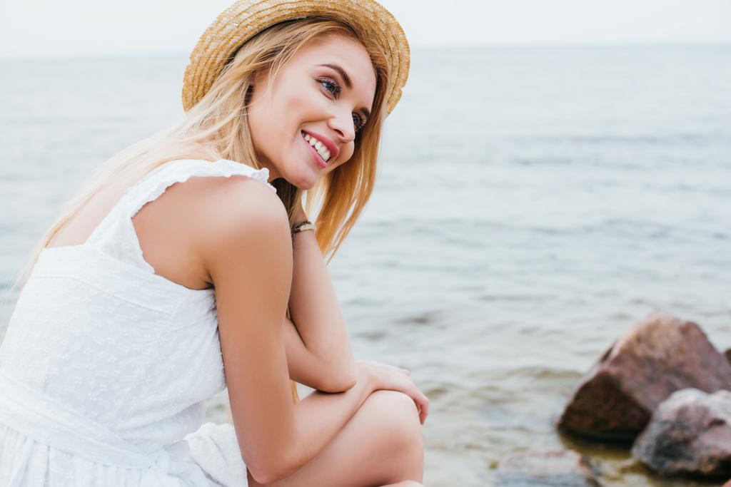 iloinen blondi nainen valkoisessa mekossa ja olkihattu hymyilee lähellä merta
  - Valokuva, kuva