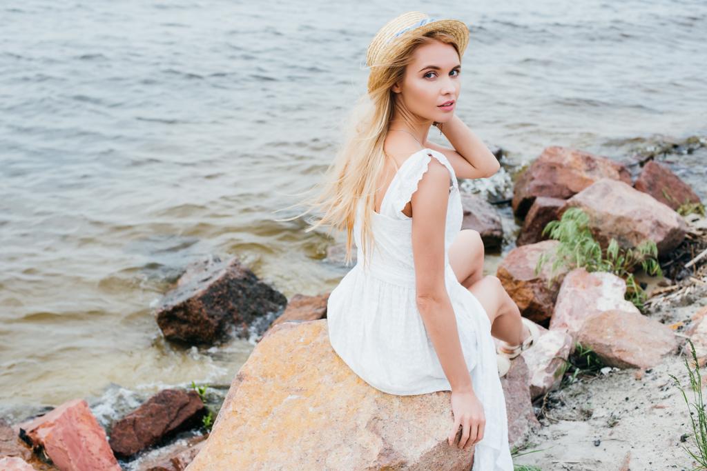 mooie blonde meisje aanraken stro hoed terwijl zittend op stenen in de buurt van zee - Foto, afbeelding