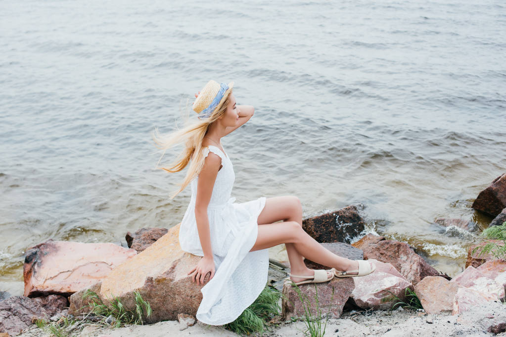 ξανθιά κορίτσι αγγίζοντας ψάθινο καπέλο, ενώ κάθονται σε πέτρες σε λευκό φόρεμα  - Φωτογραφία, εικόνα