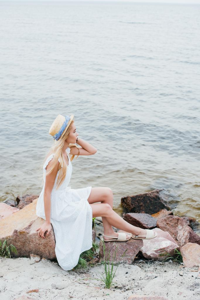 dreamy blonde girl touching straw hat while sitting on stones near sea - Photo, Image