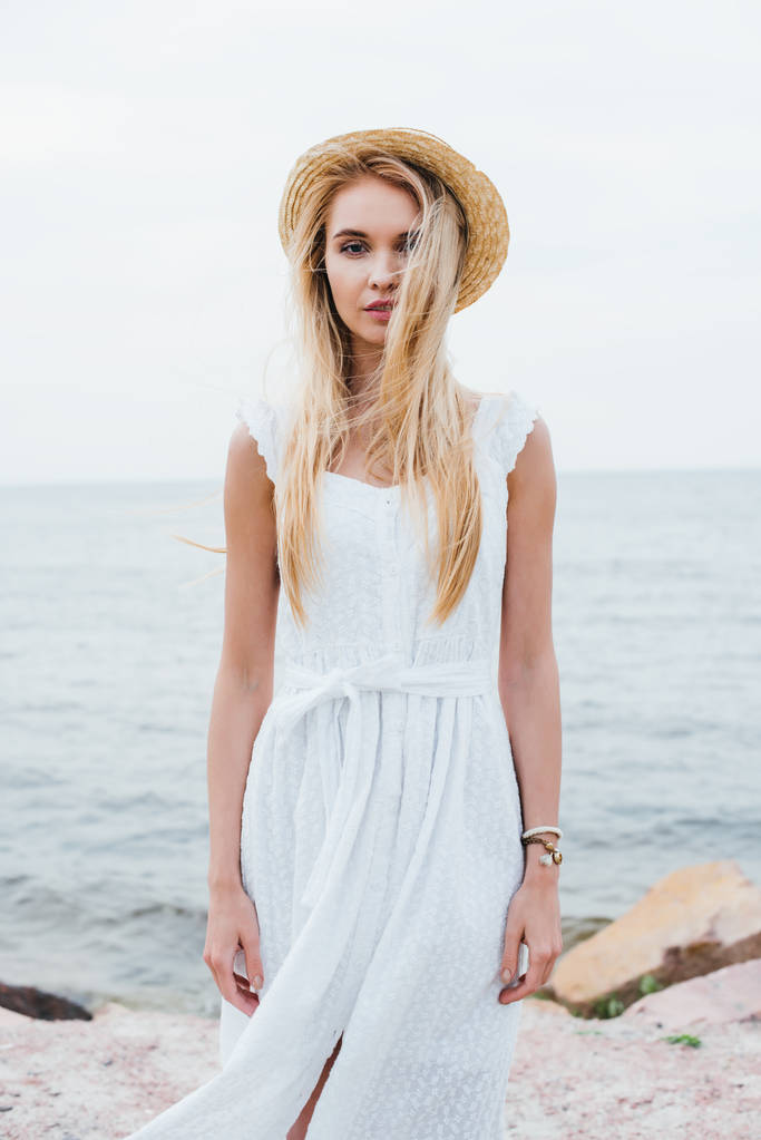 приваблива молода жінка стоїть в білій сукні і солом'яному капелюсі біля моря
  - Фото, зображення
