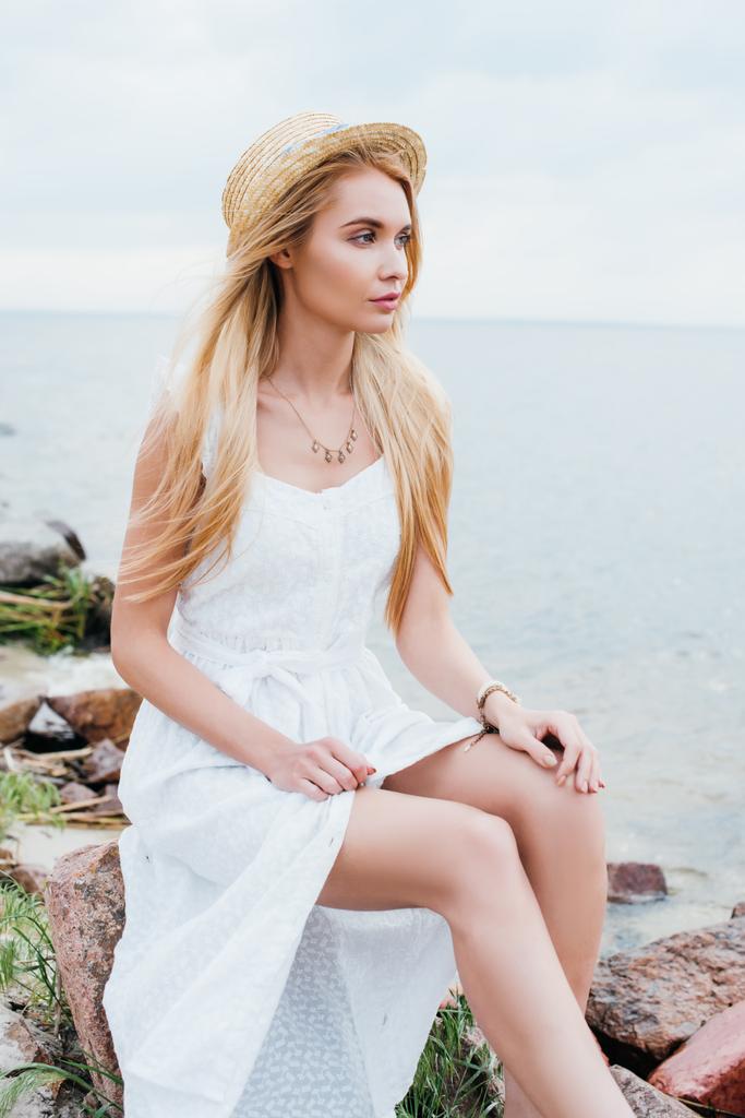 atrakcyjny blond kobieta w białej sukni i słomy kapelusz siedzi w pobliżu morza  - Zdjęcie, obraz