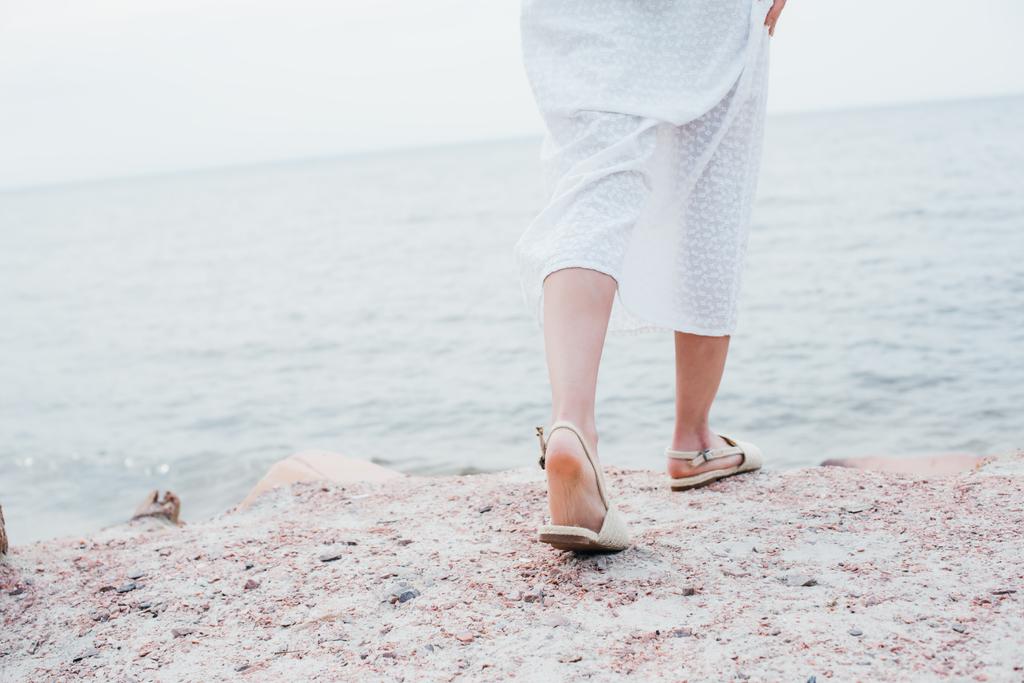 przycięte widok młodej kobiety w białej sukni i sandały stojące w pobliżu morza  - Zdjęcie, obraz