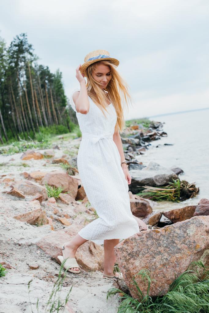 красива блондинка стоїть і торкається солом'яного капелюха біля моря
  - Фото, зображення