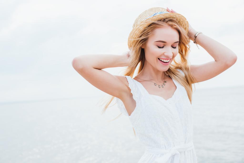 happy blonde woman with closed eyes standing and touching straw hat near sea  - Photo, Image