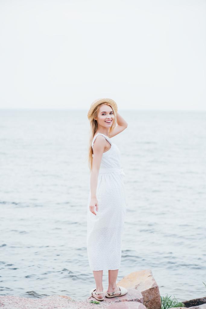 χαμογελαστή ξανθιά νεαρή γυναίκα αγγίζοντας ψάθινο καπέλο και κοιτάζοντας την κάμερα, ενώ στέκεται σε λευκό φόρεμα κοντά στη θάλασσα  - Φωτογραφία, εικόνα