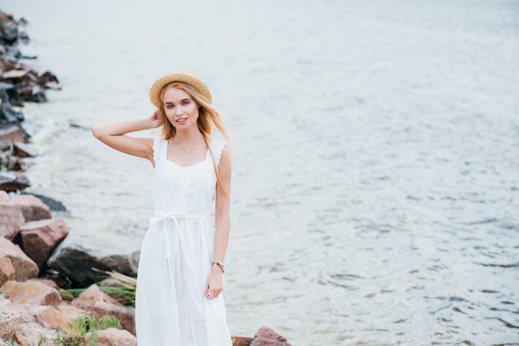 Veselá Mladá blondýnka dotek slaměných klobouků a pohled na kameru blízko moře  - Fotografie, Obrázek